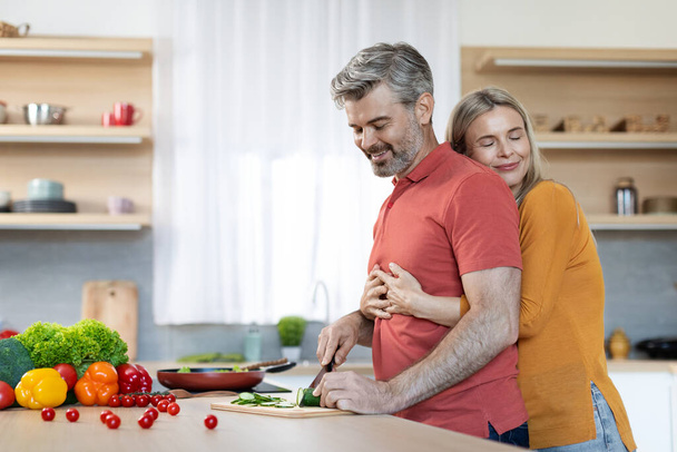 Loving handsome middle aged man husband preparing delicious healthy meal for his wife, happy blonde lady hugging her spouse cooking dinner, kitchen interior, side view, copy space - Fotoğraf, Görsel