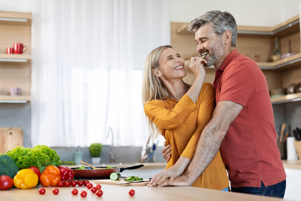 Happy middle aged spouses preparing delicious dinner together, cheerful attractive blonde woman feeding her handsome husband while cooking at home, having fun, kitchen interior, copy space - Фото, изображение