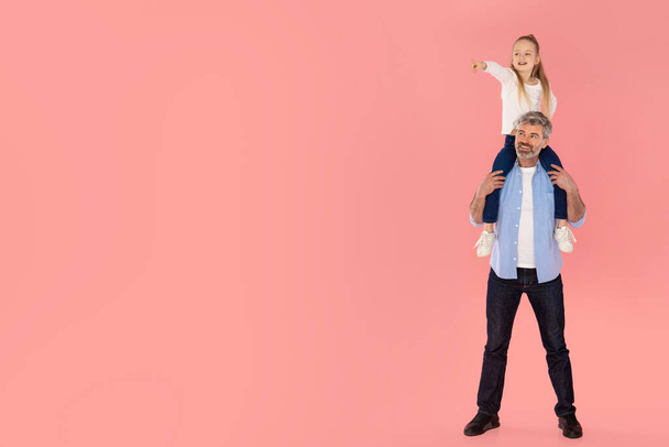 Little Daughter Pointing Finger Aside Sitting On Fathers Shoulders Advertising Family Offer Together Over Pink Studio Background. Look There Concept. Full Length, Empty Space For Text - Valokuva, kuva