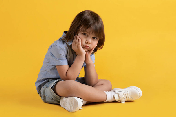 Kids loneliness. Sad unhappy little boy sitting on floor and suffering alone, leaning his chin on on hands, looking sadly at camera, yellow studio background, free space - 写真・画像
