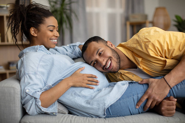 Pregnancy Lifestyle. Loving Black Spouses Hugging, Husband Listening To Pregnant Wifes Belly Smiling Expecting Baby At Home. Young Parents Enjoying Expectation. Family And Childbirth - Фото, изображение