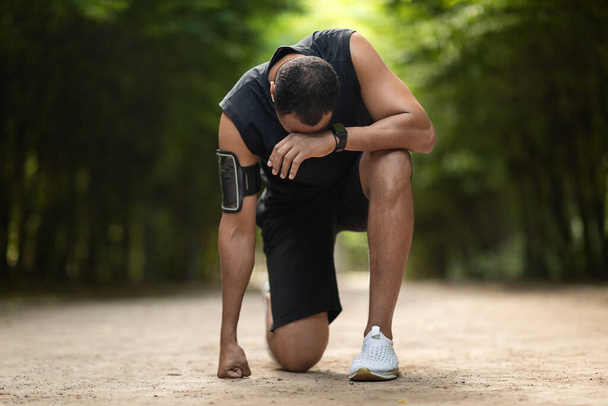 Black sportsman feeling bad while jogging by public park, african american guy in sportswear standing on his knee, touching head, got exhausted during workout outdoors, copy space - Photo, image