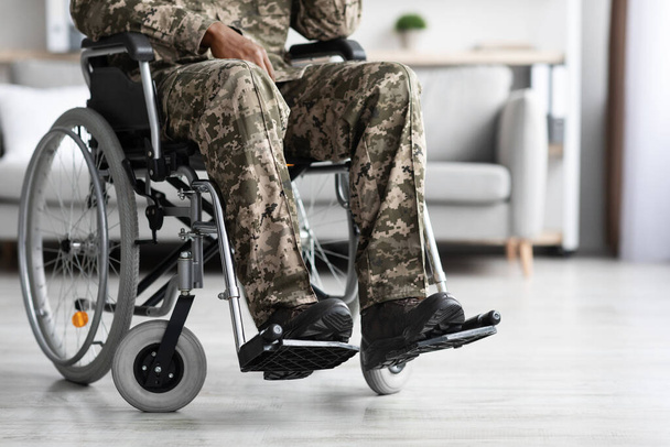 Unrecognizable black man sitting in wheelchair, wearing military uniform, suffering from loneliness and posttraumatic stress and physical disorder, home interior, panorama with copy space, cropped - Foto, Imagen