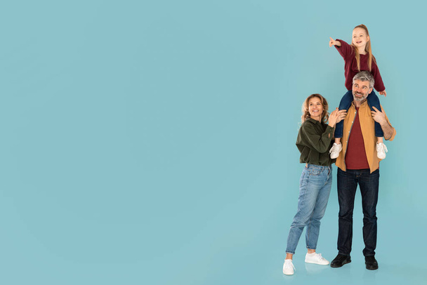 Family Offer. Kid Girl Pointing Finger Aside Sitting On Fathers Shoulders Posing With Parents Over Blue Studio Background. Full Length Shot, Empty Space For Text Advertisement - Foto, immagini