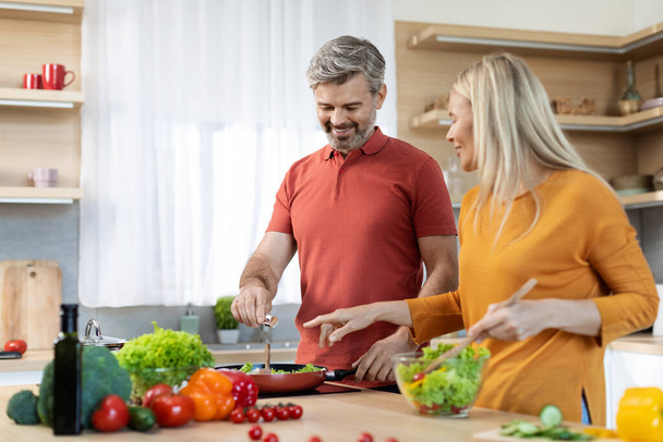 Cheerful beautiful family middle aged husband and wife enjoying cooking together at home, happy man and woman standing next to stove, preparing healthy dinner and chatting, copy space - Fotó, kép