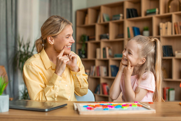 Female speech-language pathologist having lesson with little girl, teacher and pupil working on pronunciation, touching faces and grimacing, sitting at desk - Zdjęcie, obraz