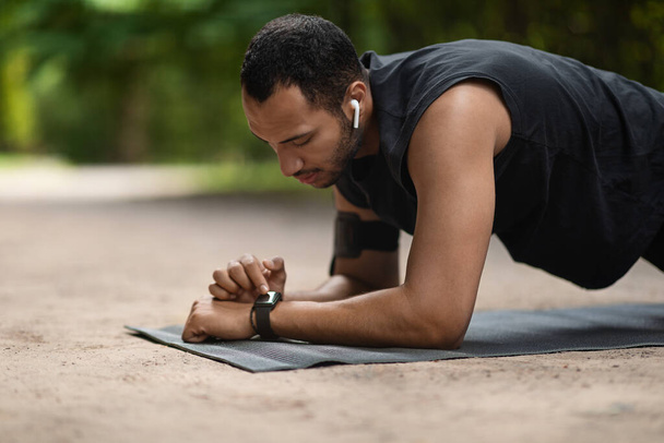 Closeup of handsome young sporty black man planking at public park, using fitness mat, setting fitness tracker, side view, copy space. Modern technologies and sport concept - Фото, изображение