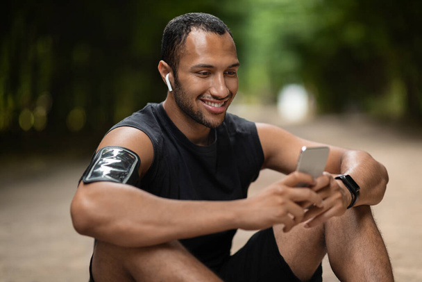 Happy handsome millennial black guy sportsman sitting on the ground, having break while training in public park, using modern smartphone and white earbuds, checking workout results, copy space - Φωτογραφία, εικόνα