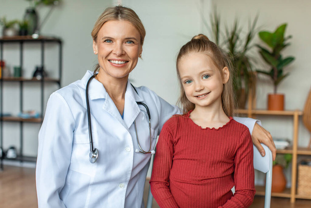 Портрет щасливого педіатра і гарненької дівчинки, яка дивиться і посміхається на камеру, сидячи в кабінеті лікарів після огляду дітей. Концепція професійних дітей
 - Фото, зображення
