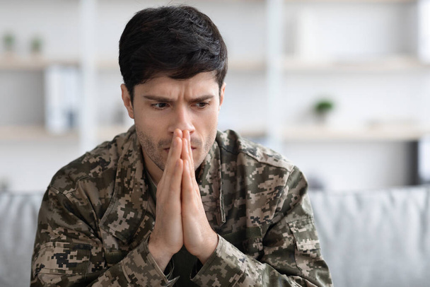 Thoughtful military man staring aside, holding palms by mouth, sitting on couch at home. Young soldier visiting psychologist, suffering from posttraumatic stress, closeup photo, copy space - Foto, Imagen