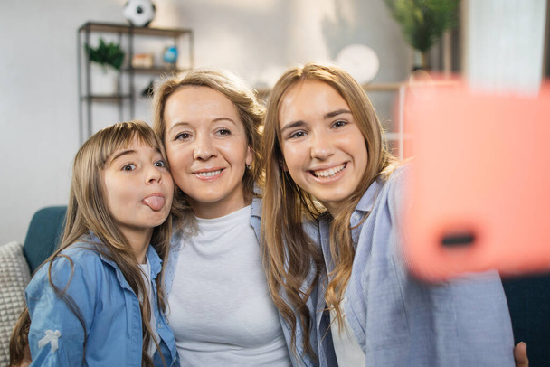 Front view happy pretty mommy with two cuddling small kids daughters, taking selfie shot on smartphone or playing online game at home. Loving young mother embracing her children, using cell phone. - Foto, Imagen