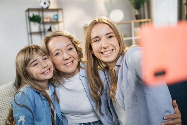 Front view happy pretty mommy with two cuddling small kids daughters, taking selfie shot on smartphone or playing online game at home. Loving young mother embracing her children, using cell phone. - Фото, изображение