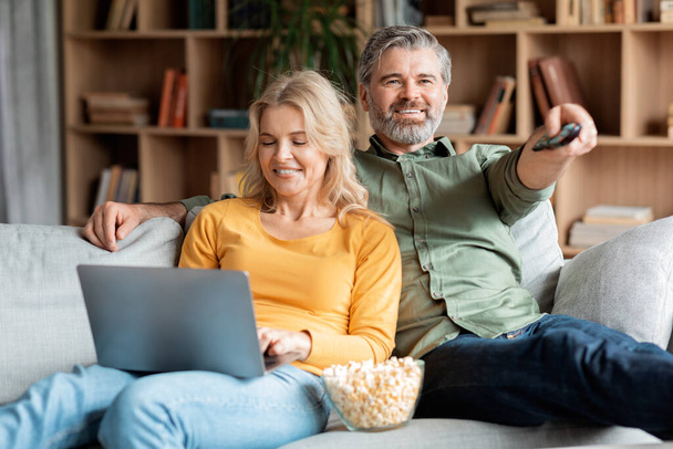 Happy Mature Couple Resting On Couch At Home, Man Watching Tv And Woman Using Laptop, Happy Middle Aged Spouses Spending Time Together In Living Room, Enjoying Domestic Leisure, Closeup - Photo, image
