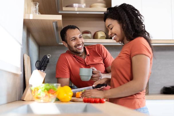 Cheerful young black couple in same t-shirts talking, wife preparing salad, man drink coffee in cozy kitchen interior. Cooking together, love and relationship, coffee break and healthy food at home - Fotoğraf, Görsel