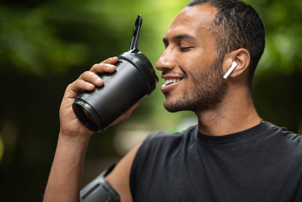 Closeup of handsome young african american man drinking protein cocktail, training outdoors, using wireless earbuds, listening to music while jogging by forest, copy space. Sports nutrition concept - Foto, immagini