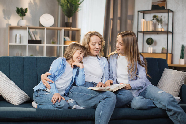 Happy loving family at home. Pretty young caucasian mother reading a book to her daughters sitting on couch in bright living room. Free time educative activity. - Foto, Bild