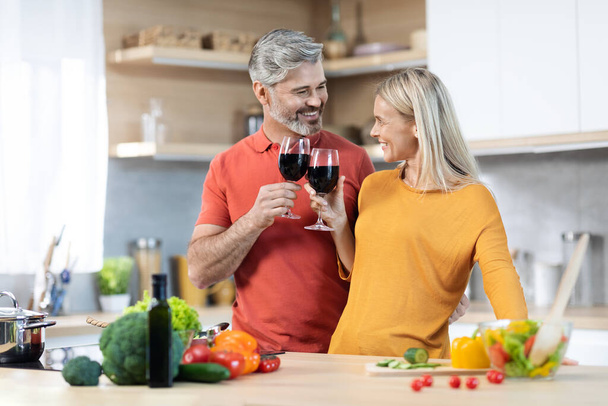 Happy middle aged couple drinking red wine while cooking, man and woman in casual standing by kitchen table, cheering with glasses, enjoying time together, celebrating anniversary, copy space - Valokuva, kuva