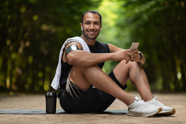 Cheerful attractive african american guy jogger sitting on yoga mat with towel on his shoulder, holding cell phone, using white earbuds, drinking protein cocktail, having break while training outdoors - Foto, Bild