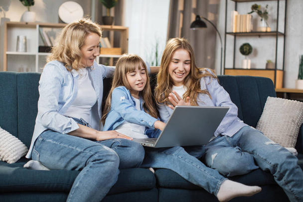 Happy family of mother and two cute daughters sitting together on comfortable couch, using laptop, resting and spend free time at home, recording video in living room. - 写真・画像