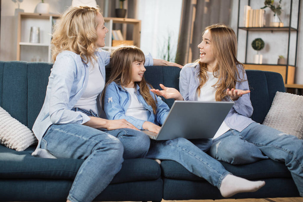 Happy family with two children having good time using laptop. Young smiling mother with cute blond daughters sitting on sofa at home looking at computer screen and watching video. - Foto, Bild