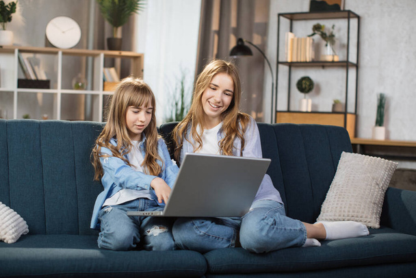 Happy sisters or friends two cute blond girls sitting together on comfortable couch, using laptop, holding video call with father or greeting grandparents, recording video in living room. - Фото, зображення