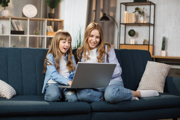 Happy sisters or friends two cute blond girls sitting together on comfortable couch, using laptop, holding video call with father or greeting grandparents, recording video in living room. - Foto, imagen