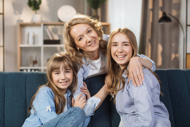 Portrait of happy two daughters hugging pleasant smiling middle age mother, relaxing together on couch. Affectionate two generations family looking at camera, posing for photo at home. - Fotó, kép
