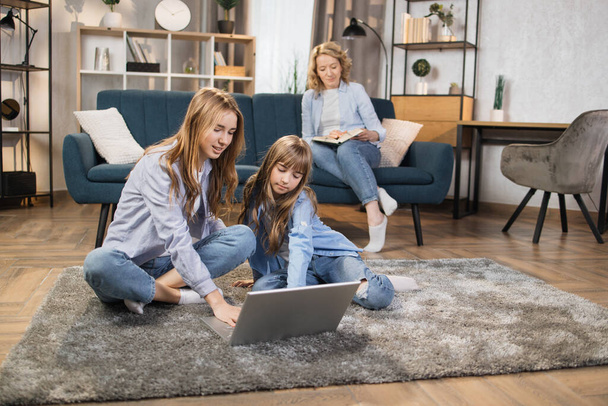 Cute blond girls sisters sitting on warm floor play at home together using laptop, while happy mother relax on sofa on background. - 写真・画像
