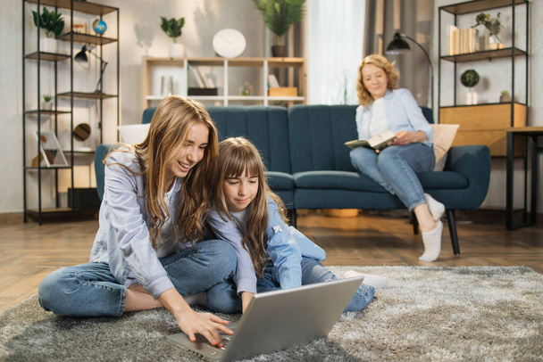 Cute blond girls sisters sitting on warm floor play at home together using laptop, while happy mother relax on sofa on background. - Photo, Image