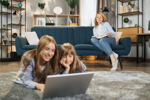 Family, leisure and people concept. Focus on happy mother with book sitting on sofa and looking at her beautiful blond daughters lying on carpet with laptop on floor at home. - Foto, Imagen