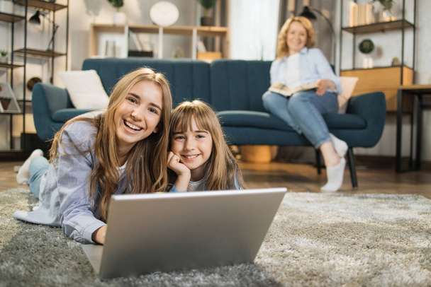 Family, leisure and people concept. Focus on happy mother with book sitting on sofa and looking at her beautiful blond daughters lying on carpet with laptop on floor at home. - Foto, imagen