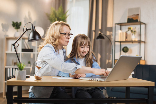 Caucasian young blond woman mother help her beautiful daughter to do homework using laptop computer sitting on table in living room at home. - Foto, immagini