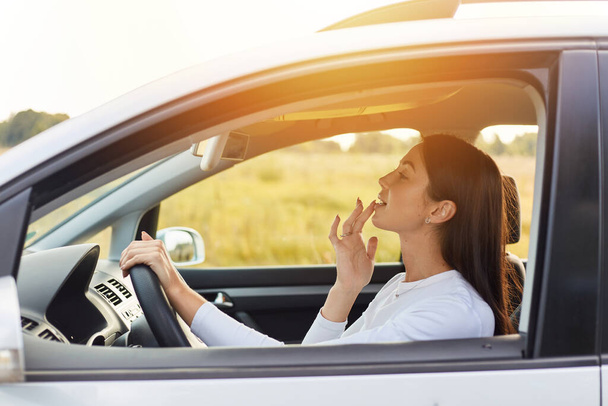 Side view portrait of dark haired beautiful woman driving a car and looking at mirror touching up her makeup, attractive female traveling alone on sunset. - Photo, image