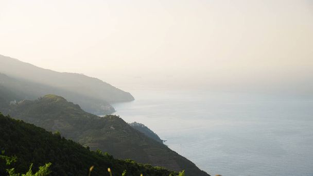 View of Ligurian sea coast, Italy. A lot of greenery, sunlight and fog, rocky coast, few buildings in the distance - Foto, imagen