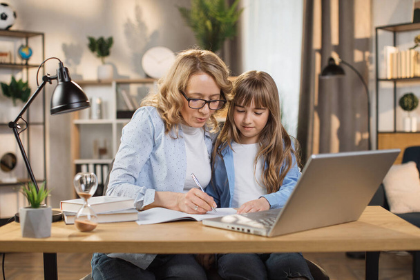 Caucasian tutor engaged with schoolgirl help complete tasks sitting at table at home. Concept of education child development, little child homeschooling writing homework with stepmother. - Foto, afbeelding