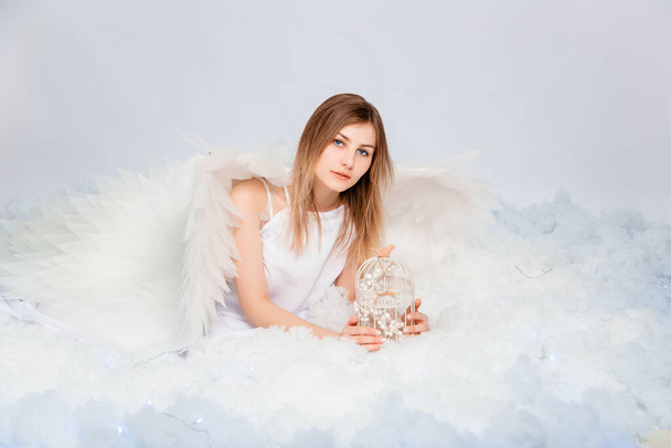 An attractive girl in a white tunic with large white wings behind her back poses while lying in white luminous clouds. - Foto, afbeelding