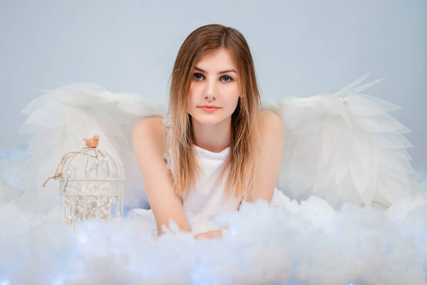 An attractive girl in a white tunic with large white wings behind her back poses while lying in white luminous clouds. - Fotó, kép