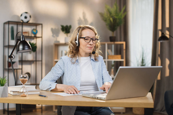 Portrait of beautiful confident with blonde hair and on headphones clever woman wearing casual clothes, sitting at table in front of computer and checking information in presentation on internet - 写真・画像