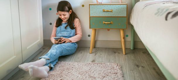 Маленькая девочка смеется, глядя на мобильный телефон, сидящий на полу в ее спальне - Фото, изображение