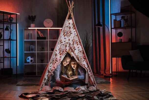 Happy blond woman nanny or older sister reading book to her lovely younger sister inside teepee tent. Comfort at home, evening storytelling and family concept. - Foto, Imagem