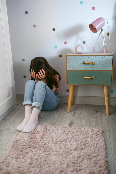 Невизначена пригнічена маленька дівчинка, що закриває обличчя руками, сидячи на підлозі у своїй спальні
 - Фото, зображення