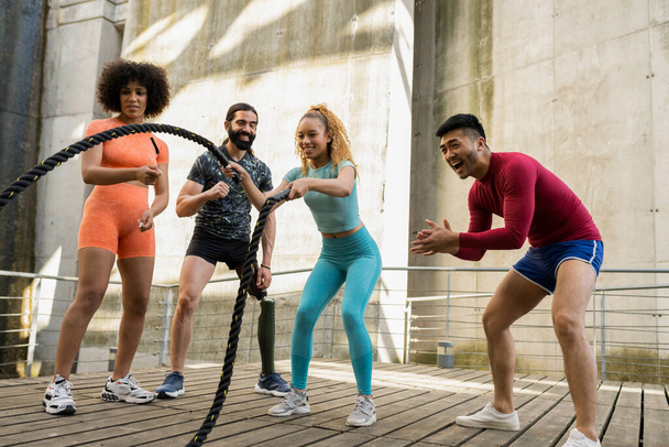 Fitness multiracial people exercising with battle ropes at the gym. Hispanic woman using the ropes training alongside her peers who cheer her on, arms and cardio. - Photo, image