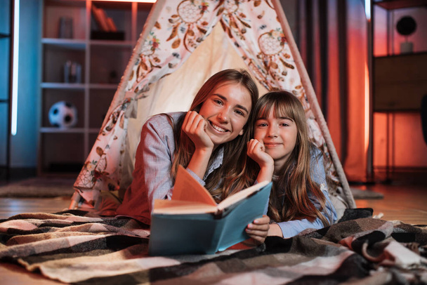 Portrait of happy caucasian loving sisters smiling and looking at camera inside wigwam. Beautiful girls and open book lying near on floor. - Fotó, kép