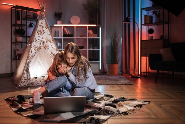 Scared blond woman, nanny or older sister hugging while watching horror movie with her lovely younger sister outside teepee tent. Evening leisure and family concept. - Foto, immagini