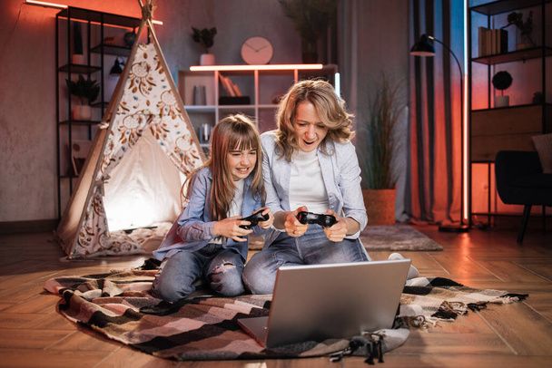 Caring caucasian senior mother or nanny with her cute daughter girl outside toy wigwam playing video games using joysticks and laptop. Dark atmosphere at cozy home. - Foto, afbeelding