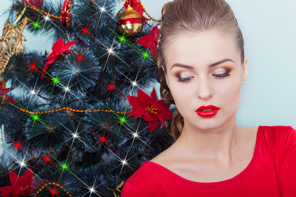 Beautiful sexy happy smiling young woman in evening dress with bright makeup with red lipstick sitting near the Christmas tree in a festive Christmas evening - Foto, Bild