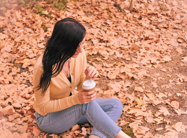 Brunette woman in yellow t-shirt snd blue jeans sitting on orange leaves in autumn and thinking about something  - Fotó, kép