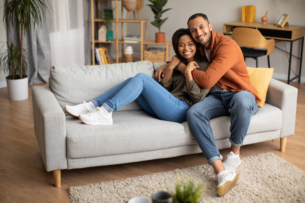 Romantic Relationship. Happy African American Spouses Hugging Smiling To Camera Sitting On Couch At Home. Husband Embracing Wife Resting On Sofa In Living Room. Marriage Concept - Valokuva, kuva