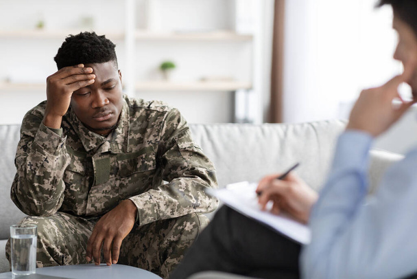 Upset african american man in military uniform veteran sitting on couch, touching his head while having conversation with psychologist, depressed soldier attending psychotherapist at clinic - Фото, изображение