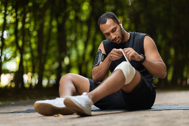 Knee injury and workout concept. African american sportsman sitting on fitness mat, wrapping injured knee with elastic bandage before training, exercising in public park, copy space - Foto, Imagem